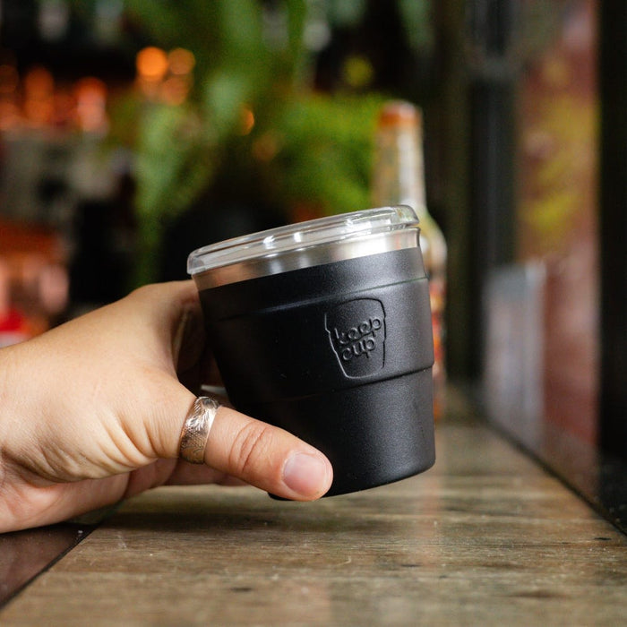 Hand holding a black KeepCup Thermal Quicksip XS 6oz reusable coffee cup with clear lid, resting on a rustic wooden table in a café setting with blurred greenery in the background.