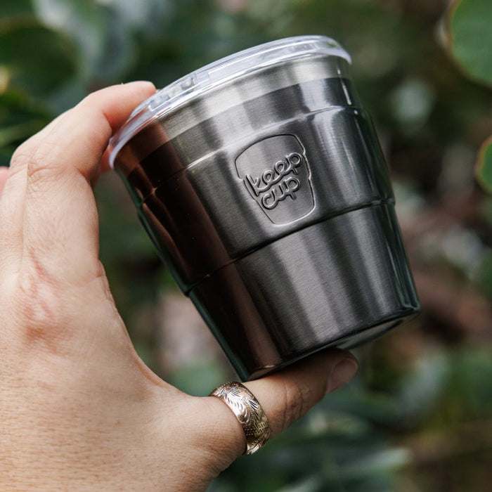 Hand holding a KeepCup Thermal Quicksip XS 6oz in Nitro, showcasing the brushed stainless steel finish with embossed KeepCup logo and clear press-fit lid, set against a soft-focus green foliage background.