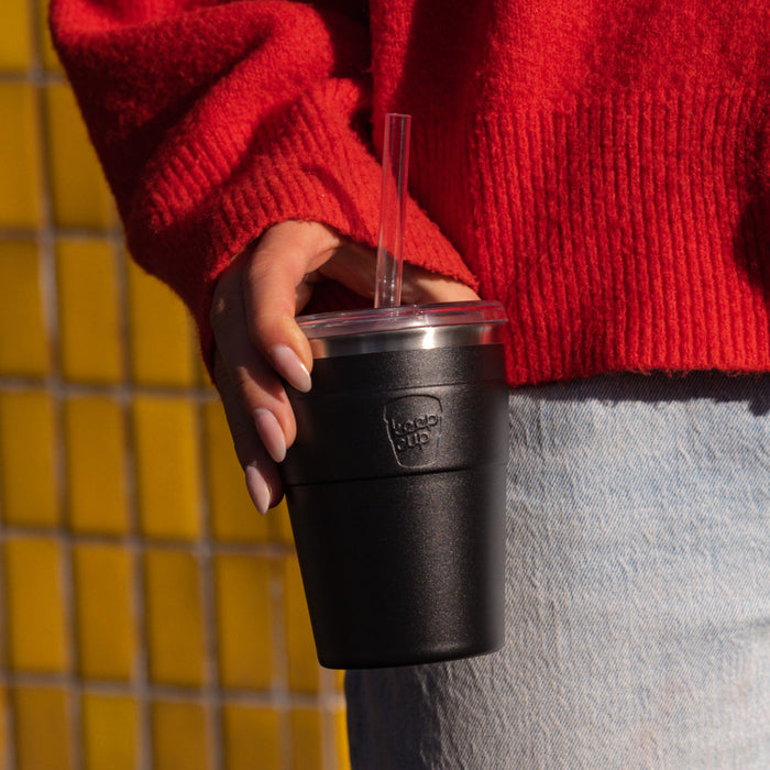 A hand with manicured nails holds a KeepCup Cold Cup Thermal M 12oz in Black at hip level, featuring a clear lid with straw, worn with a red knit jumper and light denim jeans against a yellow tiled background.