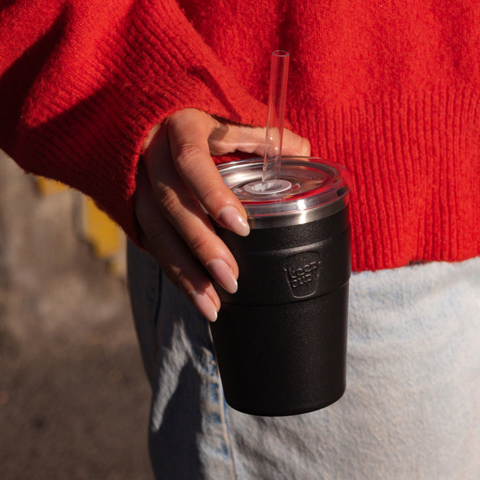 A hand with manicured nails holds a Cold Cup Thermal M 12oz in Black, featuring a clear lid with straw, against a bright red knit jumper and light denim jeans in an outdoor setting.