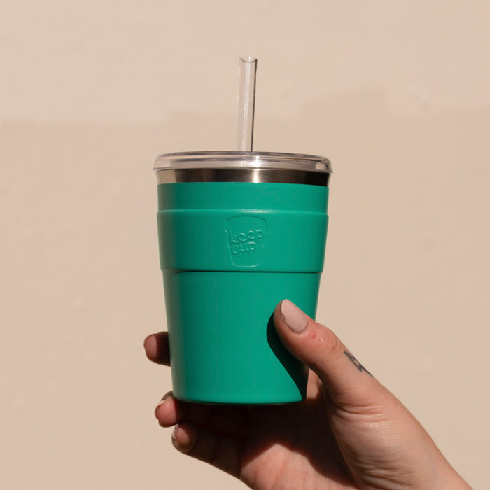 A hand with nude-coloured nail polish holds a KeepCup Cold Cup Thermal M 12oz in Frost Green, featuring a stainless steel rim, clear lid with transparent straw, and matching teal silicone band, against a neutral beige background.