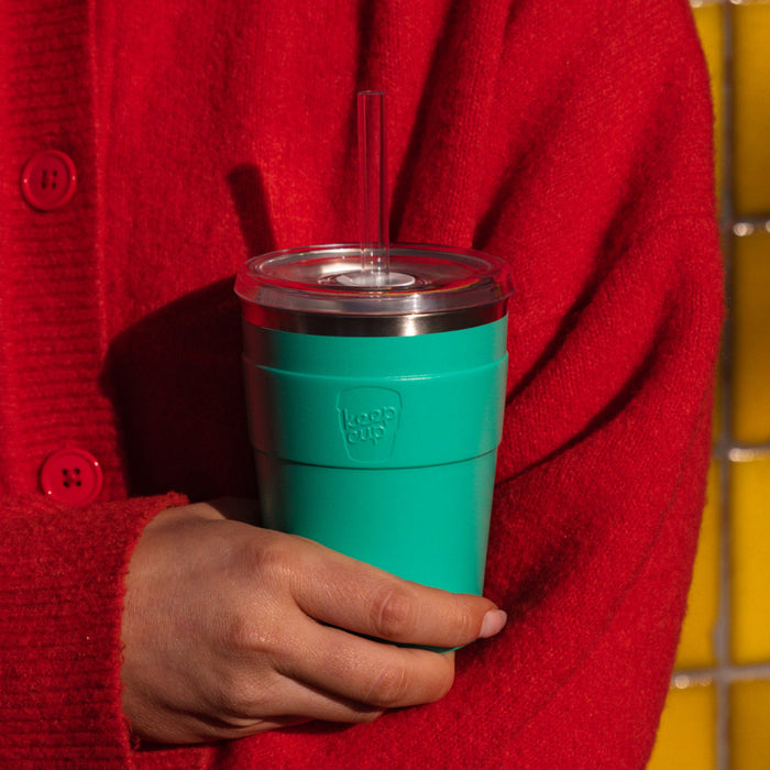 A person in a red wool cardigan cradles a KeepCup Cold Cup Thermal M 12oz in Frost Green against their body, showcasing the stainless steel tumbler with clear lid and reusable straw.