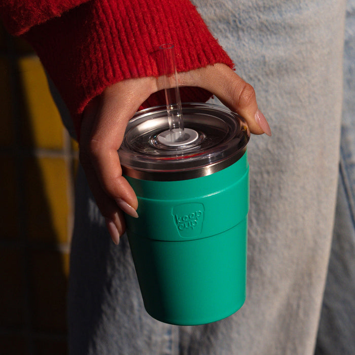 A hand with manicured nails holds a KeepCup Cold Cup Thermal M 12oz in Frost Green, featuring a clear lid with straw, against a casual backdrop of light denim jeans and a red knit jumper.