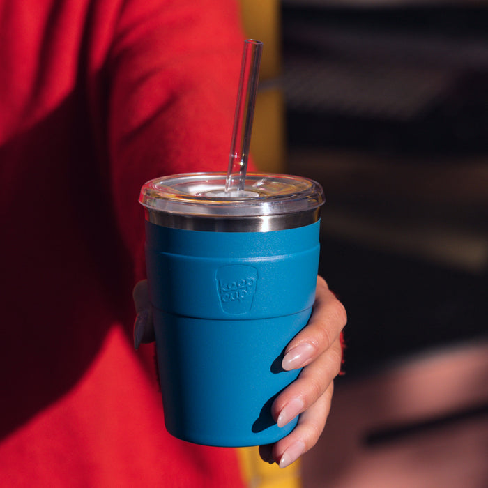 A hand with manicured nails holds a KeepCup Cold Cup Thermal M 12oz in Kingfisher blue, featuring a clear lid with reusable straw, with the person wearing a red top visible in the background.