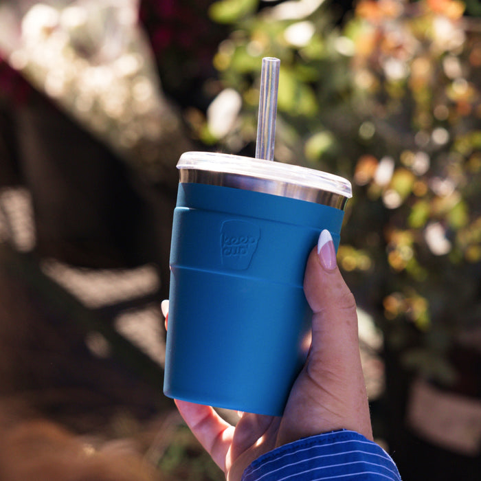 A hand with pink nail polish holds a KeepCup Cold Cup Thermal M 12oz in Kingfisher, featuring a deep teal silicone sleeve, stainless steel lid, and clear straw, with a softly blurred outdoor garden setting in the background.