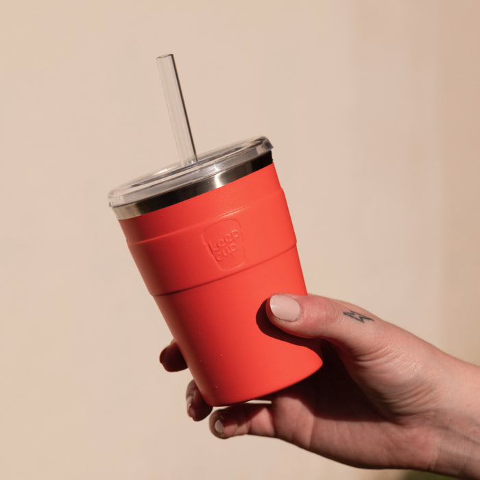 A hand with neutral nail polish holds a KeepCup Cold Cup Thermal M 12oz in Persimmon, featuring a vibrant coral-red stainless steel body with silicone grip band, clear lid and reusable straw, photographed against a soft beige background.