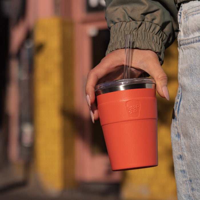 A hand with manicured nails holds the KeepCup Cold Cup Thermal M 12oz in Persimmon by the silicone band, with a clear straw visible, against an urban streetscape background with warm golden and terracotta tones.