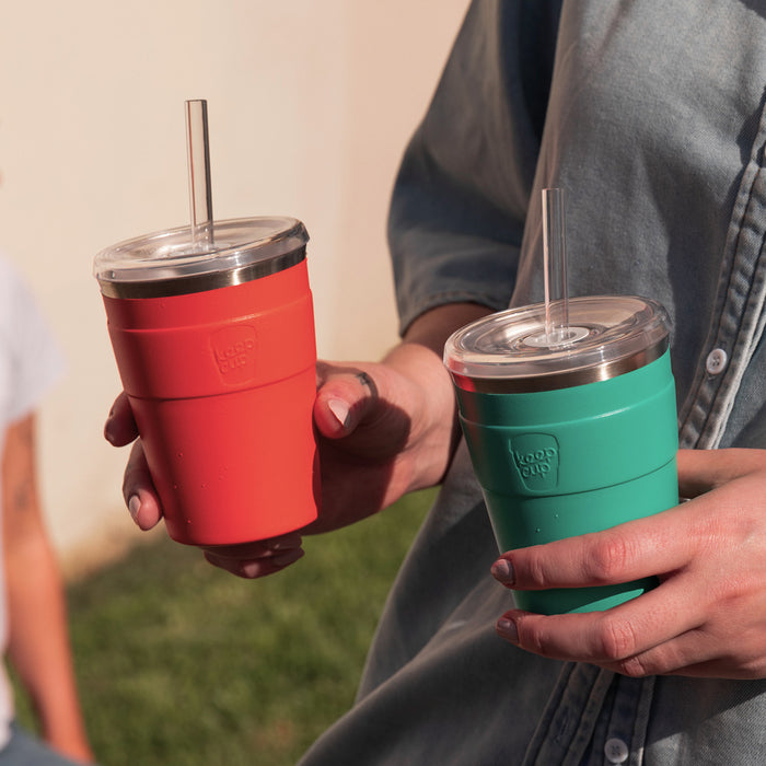 Two people holding KeepCup Cold Cup Thermals with reusable straws outdoors, featuring the M 12oz Persimmon in coral red alongside a teal colourway, both with clear lids and stainless steel rims.