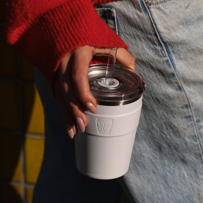 A hand with manicured nails holds a Cold Cup Thermal M 12oz in White, showing the reusable cup's clear lid with straw opening and light grey silicone grip band, against a casual backdrop of a red knit jumper and denim jeans.