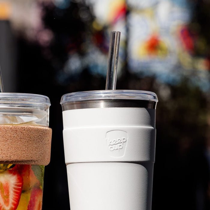 White KeepCup Cold Cup Thermal M 12oz with clear lid and stainless steel straw, shown alongside a glass KeepCup with cork band containing fruit-infused water, against a colourful blurred background.