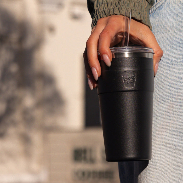 A woman's hand with manicured nails holds a black KeepCup Cold Cup Thermal L 16oz by her side, featuring a clear lid with straw, against a softly blurred urban background.