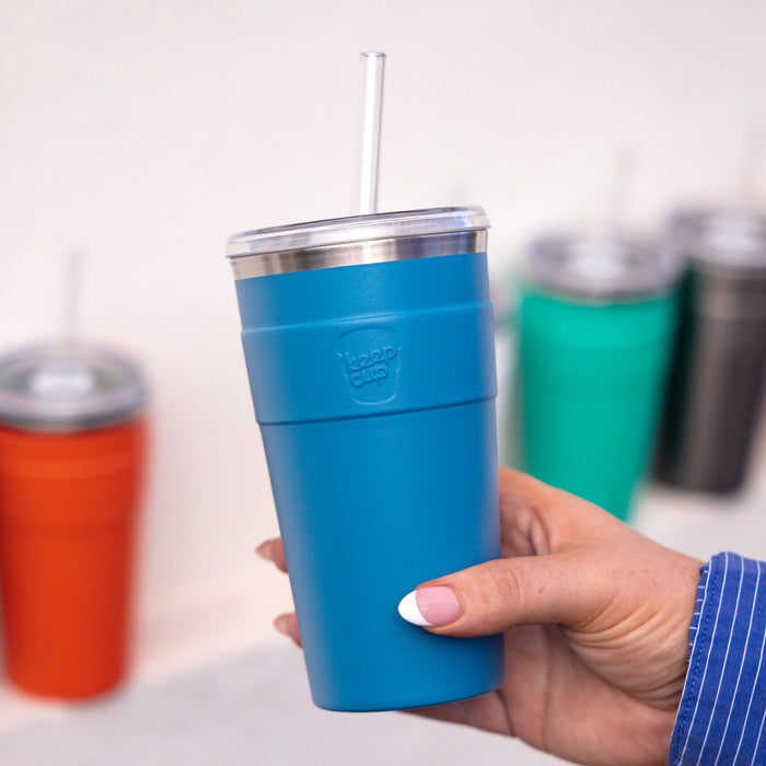 Hand holding a KeepCup Cold Cup Thermal L 16oz in Kingfisher, a matte teal blue insulated tumbler with stainless steel rim and clear straw, with orange, green and black KeepCups blurred in the background.