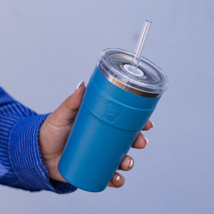 A hand with white nail polish holds the KeepCup Cold Cup Thermal L 16oz in Kingfisher, a vibrant teal blue insulated tumbler with clear lid and reusable straw, against a soft blue background.