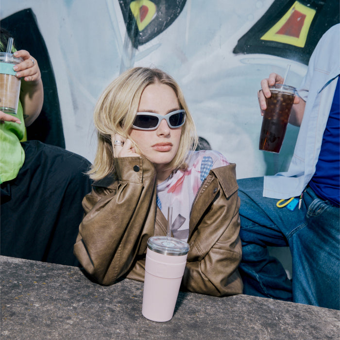 A young woman in sunglasses and a brown leather jacket leans on a concrete ledge with a KeepCup Cold Cup Thermal L 16oz in Pashmak Pink in front of her, while friends holding iced drinks sit nearby against a colourful mural backdrop.