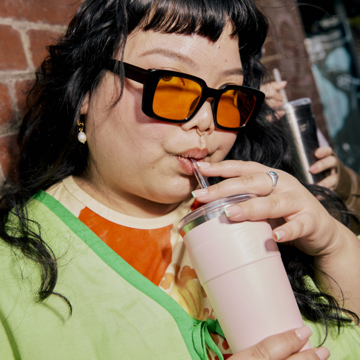 A woman with long black hair and orange-tinted sunglasses sips from a KeepCup Cold Cup Thermal L 16oz in Pashmak Pink through a clear straw, wearing a colourful green and orange outfit against a brick wall backdrop.