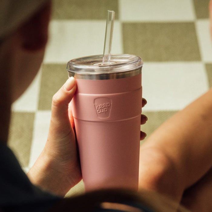 A person holds a KeepCup Cold Cup Thermal L 16oz in Pashmak Pink, viewed from over the shoulder. The reusable cup features a matte pink insulated body with embossed KeepCup logo, clear lid and straw, set against a checkered floor background.