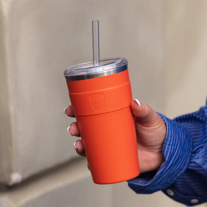 A hand with white nail polish holds a KeepCup Cold Cup Thermal L 16oz in Persimmon, featuring a vibrant orange insulated body with clear lid and reusable straw, against a neutral background.