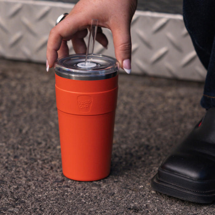 A hand with white nail polish reaches down to pick up a KeepCup Cold Cup Thermal L 16oz in Persimmon, featuring a clear lid with straw, placed on textured asphalt beside a metal diamond plate step.