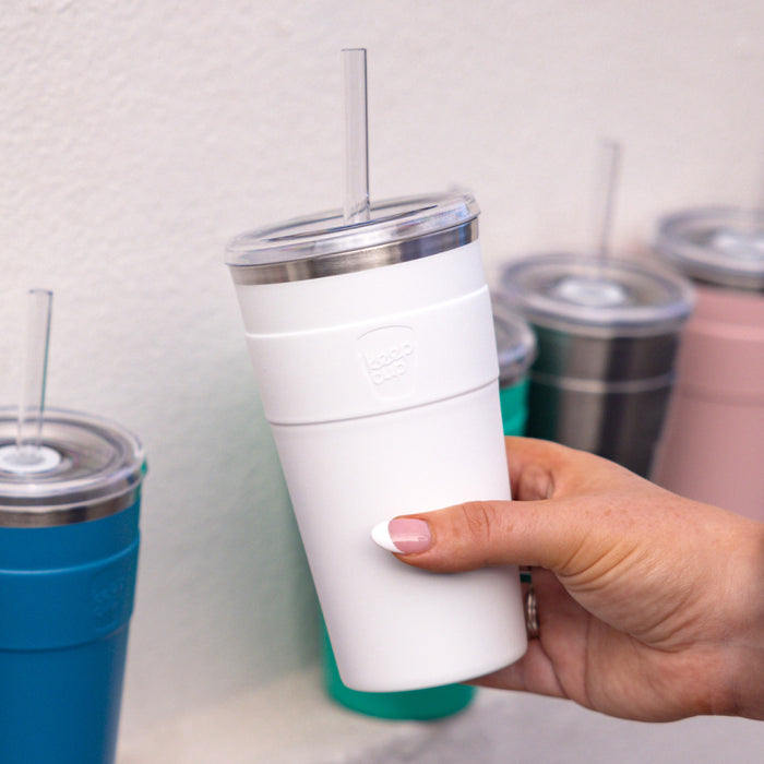 A hand with white-painted nails holds a KeepCup Cold Cup Thermal L 16oz in White, featuring a clear lid with reusable straw, with other KeepCup colours including blue, teal and pink visible on a shelf in the background.