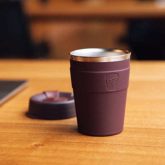 KeepCup Thermal M 12oz in Alder, a deep plum-coloured reusable cup with stainless steel rim, sitting on a wooden café table with its matching lid placed alongside.