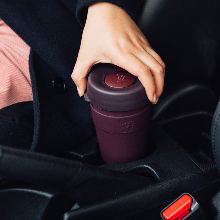 A hand reaches down to place or pick up a KeepCup Thermal M 12oz in Alder, a deep plum-coloured reusable cup, from a car cupholder in the centre console.