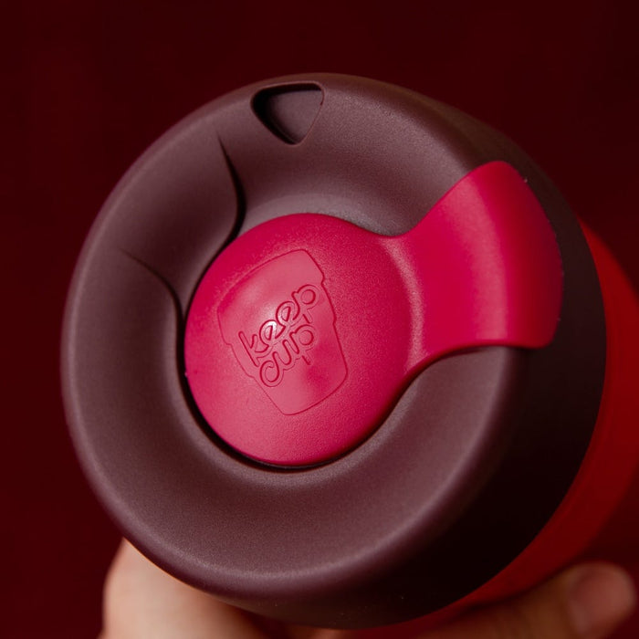 Close-up top-down view of a hand holding the KeepCup Thermal M 12oz in Ginger, showing the dark burgundy lid with the bright red sipper plug featuring the embossed KeepCup logo.