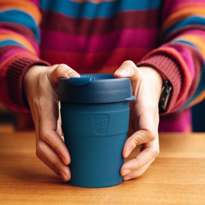 Hands cradling a KeepCup Thermal M 12oz in Lagoon, a deep teal reusable coffee cup with matching lid, resting on a wooden table with a colourful striped jumper visible in the background.