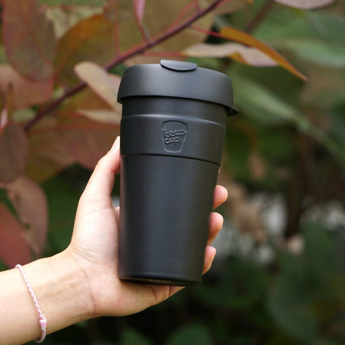 A hand holds the KeepCup Thermal L 16oz in Black, showcasing its sleek matte black stainless steel body with matching lid and embossed KeepCup logo, set against a soft-focus backdrop of green and burgundy foliage.