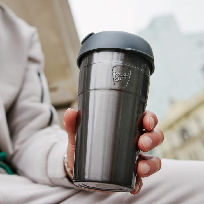 A hand holds the KeepCup Thermal L 16oz in Nitro, showcasing its brushed stainless steel body and charcoal grey lid with the KeepCup logo embossed on both, set against a blurred urban background.