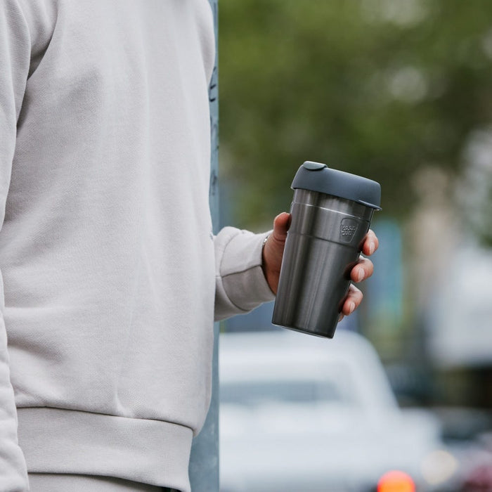 A person in a light grey sweatshirt holds a KeepCup Thermal L 16oz in Nitro, showcasing the stainless steel body and dark grey lid while standing on an urban street with blurred cars and trees in the background.