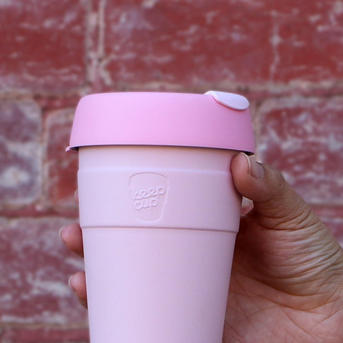 Hand holding a KeepCup Thermal L 16oz reusable coffee cup in Pashmak Pink, featuring a soft pink body with matching pink lid and white sipper plug, photographed against a weathered red brick wall.