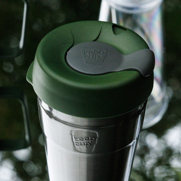 KeepCup Thermal L 16oz in Pine, featuring a stainless steel body with dark green plastic lid and grey plug, photographed at a close-up angle on a reflective surface with other reusable cups softly blurred in the background.
