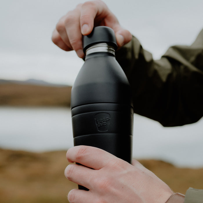 Hands opening the lid of a black KeepCup Helix Bottle 660ml outdoors, with a blurred natural landscape in the background.