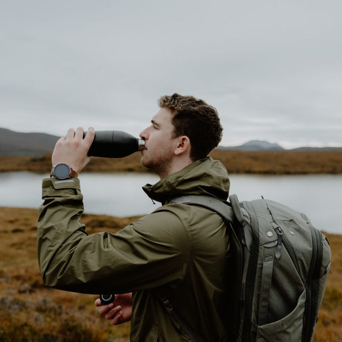A man in an olive green jacket and backpack drinks from a KeepCup Helix Bottle in Black while hiking, with a scenic lake and rugged moorland landscape in the background on an overcast day.