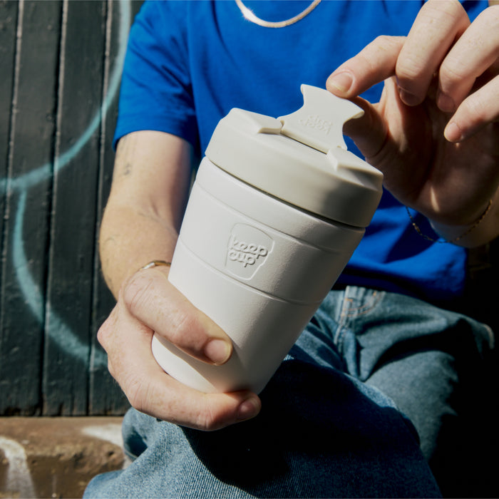 A person in a blue t-shirt sits outdoors, holding a KeepCup Commuter M 12oz in Bone White while opening the sipper lid with their other hand, showcasing the cup's embossed logo and reusable design.