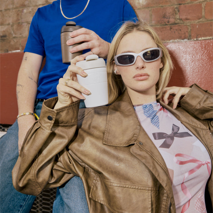 A woman in sunglasses and a brown leather jacket holds a KeepCup Commuter M 12oz in Bone White, while a person beside her in a blue t-shirt holds a Chocolate KeepCup, both relaxing against a brick wall in an urban setting.