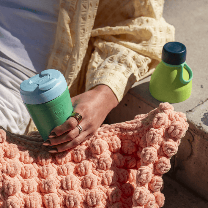 A hand wearing rings holds a KeepCup Just Essentials in Eden Cloud, featuring a green body with light blue lid, while seated outdoors on a textured peach blanket with a yellow knit cardigan visible, and a second lime green and navy KeepCup resting nearby on concrete.