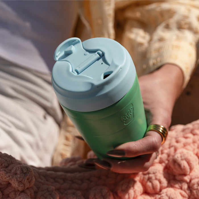 A hand wearing gold rings holds the KeepCup Just Essentials in Eden Cloud, featuring a green body and light blue lid with flip-top opening, against a cosy backdrop of textured blankets in soft pink and cream tones.