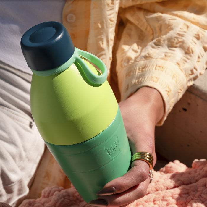 A hand with gold rings holds the KeepCup Just Essentials in Eden Cloud, featuring a lime green body, sage green base and carry loop, and dark teal lid, in a relaxed outdoor setting with soft pink and neutral tones.