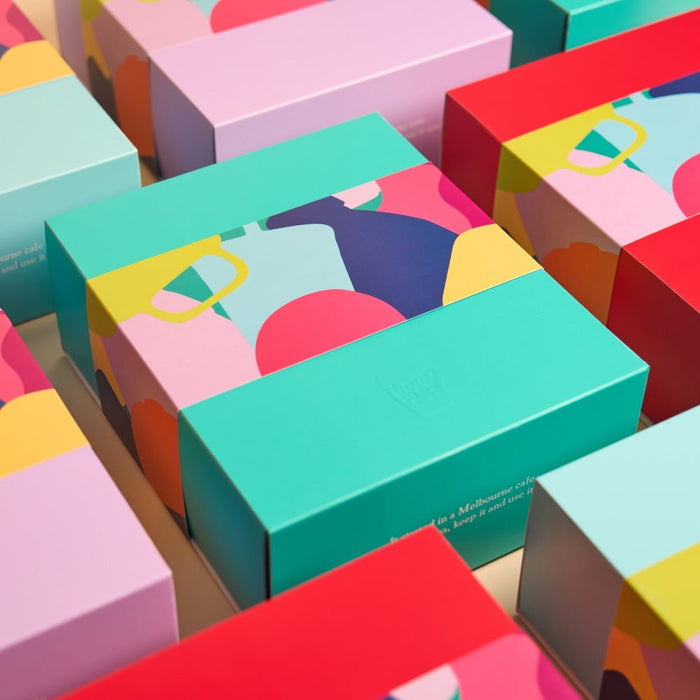 Colourful KeepCup gift boxes arranged in a grid pattern, featuring vibrant teal, pink, red and yellow packaging with abstract geometric designs and an embossed KeepCup logo, with text noting the product was designed in a Melbourne café.