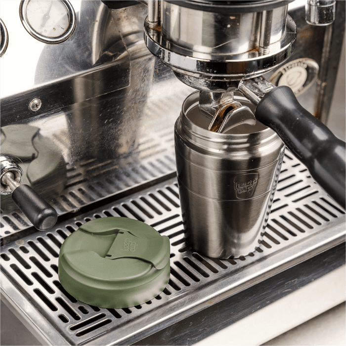 A stainless steel KeepCup Just Essentials reusable cup sits beneath an espresso machine portafilter, with espresso flowing into it, while the Pine-coloured olive green lid rests on the drip tray beside it.