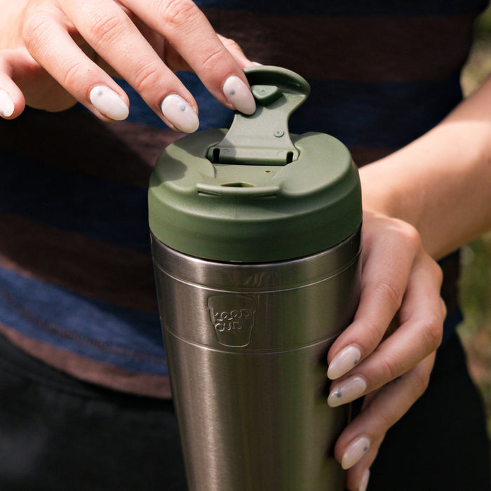 Hands with neutral nail polish opening the flip-top lid of a KeepCup Just Essentials in Pine, featuring a stainless steel body and olive green lid.
