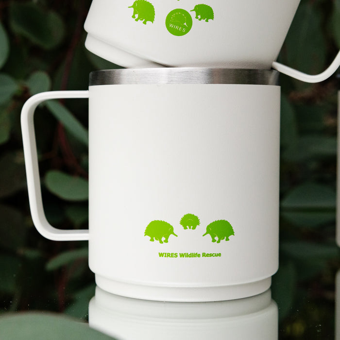 Two white KeepCup x WIRES Camp Mugs in M 12oz size stacked on top of each other, featuring lime green echidna illustrations and the WIRES Wildlife Rescue logo, photographed against a backdrop of eucalyptus leaves.