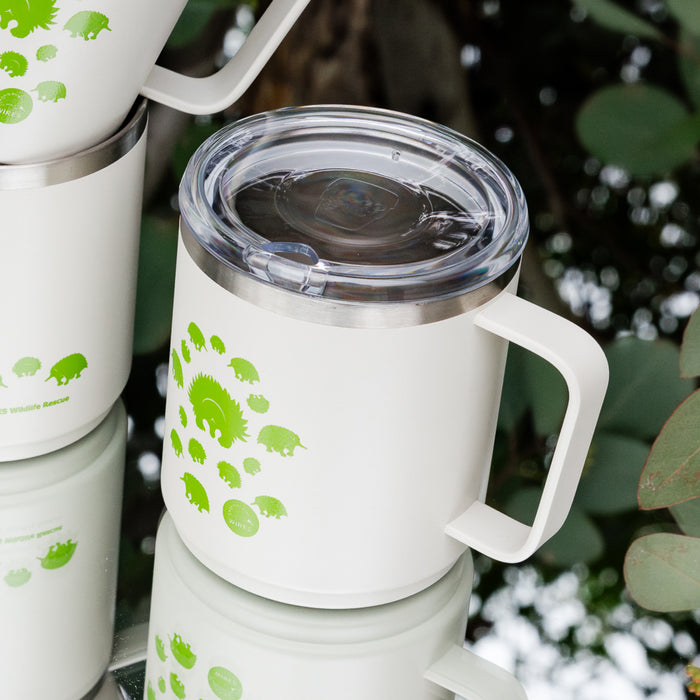 KeepCup x WIRES Camp Mug in white with green echidna illustrations arranged in a spiral pattern, featuring a clear plastic lid and sturdy handle. Multiple mugs are displayed amongst eucalyptus leaves, highlighting the wildlife rescue collaboration.