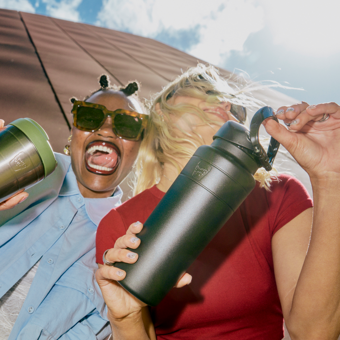 Two friends laughing and holding KeepCup Ora Bottles in a low-angle outdoor shot, with the 700ml Black bottle prominently displayed in the foreground alongside a green bottle, against a sunny blue sky.