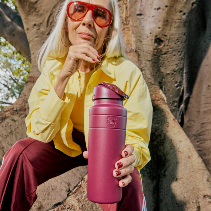 A woman with long silver hair and red sunglasses sits on a large tree trunk, holding out a KeepCup Ora Bottle in Mulberry colour toward the camera, wearing a yellow shirt and burgundy trousers with red nail polish matching the bottle.