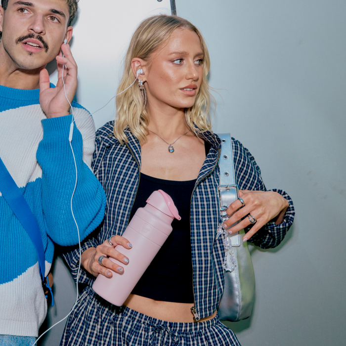 A young woman with blonde wavy hair wearing a blue checked shirt over a black top holds an Ora Bottle 700ml in Pashmak Pink while standing next to a man in a blue and white sweater, both sharing earphones against a grey backdrop.