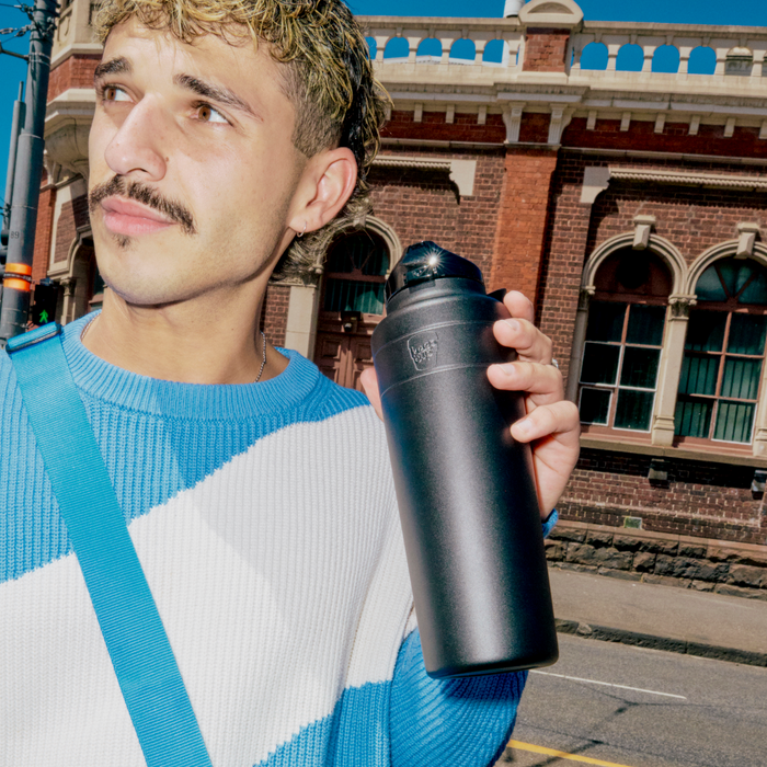A man with curly hair and a moustache wearing a blue and white knit jumper holds a black Ora Bottle 950ml while standing on an urban street with a heritage brick building in the background.