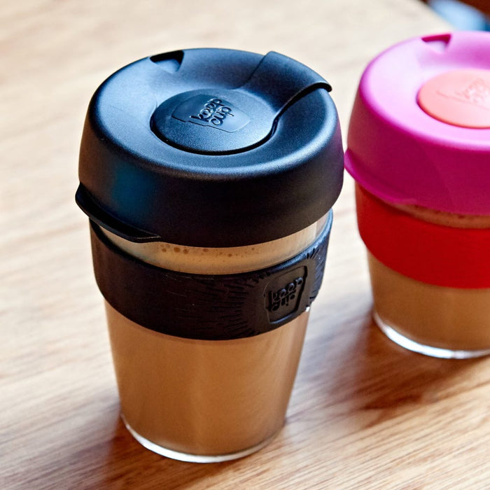 KeepCup Original Clear M 12oz in Black filled with a latte, sitting on a wooden café table alongside a pink and red KeepCup, showing the reusable plastic cup with black silicone band and lid.