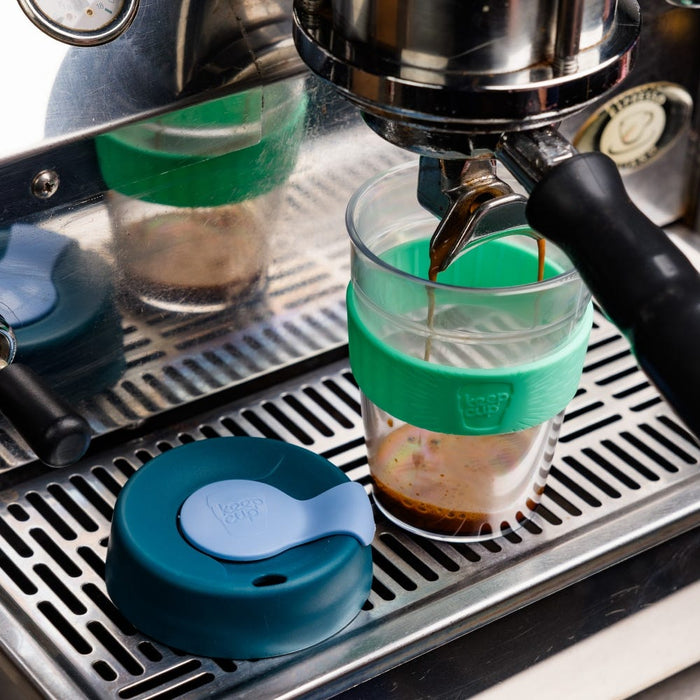 A KeepCup Original Clear M 12oz in Eventide sits on an espresso machine drip tray as coffee pours into it from the portafilter, with its teal and light blue lid placed beside it.